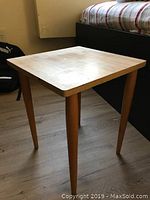 Wooden square-top stool with tapered legs placed indoors near a bed and chair.
