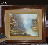 Framed oil on canvas landscape painting showing a river with trees and mountains in the background, in a wooden frame with beige matting.