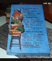 Top view showing painted blue background with hand-lettered classic literary titles and author names alongside a small metal chair supporting a stacked arrangement of miniature 3D books.