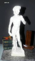 Full view of white alabaster statue of David standing on marble base beside books and small objects for scale, lit against dark background.