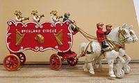 Side view of red Overland Circus bandwagon pulled by two white horses with riders in red and black attire, showing detailed gold chains and paintwork.