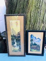 Pair of two framed watercolor landscape paintings leaning against a tree, one tall and narrow, the other smaller and horizontal.