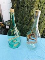 Two vintage glass bottles with folk art wooden figures inside placed outdoors on blue surface against tree background