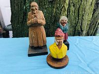 Three wooden carved figurines arranged on a blue cloth with tree bark background. Left is a tall woman in natural wood color, middle is a bust of man with red hat and yellow shirt, right is an elderly woman in blue and red clothing.