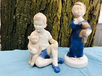 Two porcelain figurines shown on blue cloth in front of tree bark background: a boy seated holding a monkey plush and a girl standing with a teddy bear.