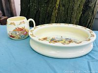 Image showing a handled children's mug alongside a shallow oval dish. Both pieces have Bunnykins bunny illustrations and border patterns.