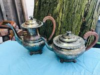 Photo showing full view of both antique silver plated tea and coffee pots with engraved floral patterns, wooden handles and clear glass finials on lids, placed on a blue cloth outdoors against a tree trunk.
