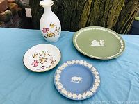 Four ceramic pieces arranged on blue cloth against backdrop of tree trunk—one vase and three plates (white floral decorated, green jasperware, blue jasperware).