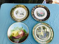 Four vintage cabinet plates on blue cloth showing unique hand-painted designs and decorative borders.