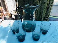 Full set of one glass pitcher and four matching glasses in smoky blue-green glass, displayed outdoors by a tree.