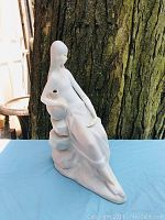Porcelain figurine of a seated woman, shown from the front and side on a blue cloth background against a tree.