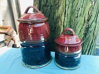 Two vintage covered pottery jars with red to dark blue glaze, lids with loop handles, placed on blue surface outdoors near a tree.