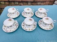 Six Royal Albert fine bone china cups and matching saucers arranged on blue tablecloth showcasing Winsome floral pattern with gold trim.