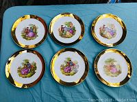 Six Limoges porcelain plates arranged on blue fabric, all with gold rims and framed Fragonard-style romantic scenes showing couples in 18th-century dress.