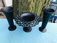 Photo of two tall black satin glass vases and a black satin glass compote bowl with open lattice rim, displayed outdoors on a blue cloth with a tree trunk background.