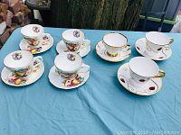 Eight porcelain cups and saucers arranged on a table showing both fruit motif and abstract gold designs.