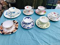 Seven pairs of English fine bone china cups and saucers arranged on outdoor table, showing varied floral and geometric patterns and colors including blue, pink, red, green and gold accents.