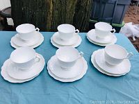 Overview of six white bone china cup and saucer trios laid out on a blue cloth, showing cups, small saucers, and larger plates with embossed pattern.