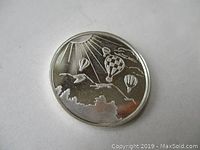 Obverse of silver coin showing hot air balloons under sun rays and clouds.