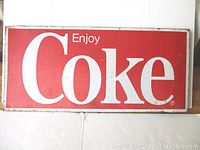 Full front view of rectangular red metal sign with white 'Enjoy Coke' text, displaying general wear and rust spots.