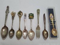 Photo of nine assorted souvenir spoons and one fork arranged in a row on white background. Various decorations including figures, emblems, and pictorial scenes.