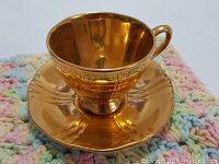 Close-up view of the golden small tea cup and saucer showing reflective metallic gold finish, textured edges, and cup handle.