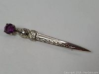 Close-up photo of a silver-toned kilt pin with an amethyst stone on one end and etched detail along its length.