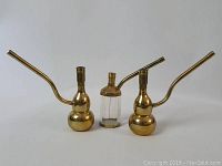 Two gold toned hookah pipes flanking a clear glass bottle with gold toned top, displayed on white background.