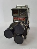 Front and angled view of Holiday II movie camera showing three-lens turret and front details with the 'Holiday II' branding below the viewfinder.