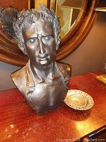 Photo showing bronze bust sculpture and small decorative metal dish together on wooden surface with mirror background.
