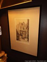 Framed black and white etching of a narrow street view labeled as Strand Lane, London, shown hanging on a dark wall with some illumination highlighting the artwork.