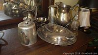 Photo showing an assortment of silver plated serving pieces including teapot, pitchers, covered serving tray, small bowl, and crystal bell arranged on a wooden surface.