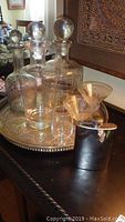 Overall view of tray with three decanters, cocktail glasses, small glass, and flask arranged on silver tray with ornate edges