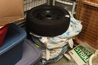 Stack of 4 tires with one tire and rim visible on top, stored near plastic bins and cardboard box.