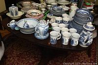 Wide shot of the entire lot on a table showing two teapots, multiple blue willow patterned mugs, bowls, plates in blue and pink patterns, and small ceramic items.