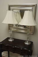 Pair of lucite and metal table lamps with white fabric shades shown on a dark wood table with a metallic framed mirror behind