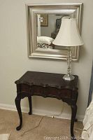 Dark stained wooden side table with one drawer, scalloped front edges, curved legs, table lamp and silver framed mirror on wall above table.