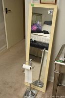 Full view of the hanging door mirror with beige plastic frame sized 51x15 inches alongside the chrome metal toilet paper stand standing next to it.