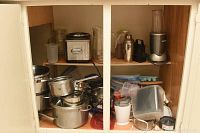 Photo inside cabinet showing an assortment of pots and pans stacked and various small kitchen appliances including a blender, toaster, coffee grinder, plastic containers, and barware.