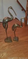 Two bronze stork sculptures standing on a wooden floor next to a staircase.