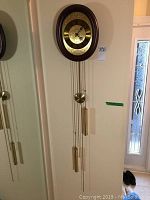Full view of the vintage Schmeckenbecher pendulum wall clock mounted on wall, showing wood frame, brass dial, pendulum, and weights hanging below.