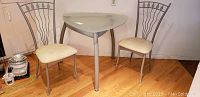 Kitchen triangle glass table with metal legs and 2 metal frame chairs with cream cushioned seats in room with hardwood floor.