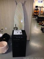 Photo of black Saviair 5000 air cleaner, ironing board with striped cover, and fireplace tools on a floor against a wall.
