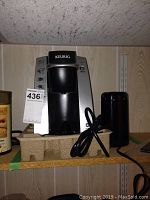 Front view of black and silver Keurig coffee maker with power cord coiled beside it on shelf.