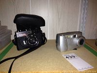Konica Auto S3 film camera in leather case and Kodak DX3215 digital camera shown side by side on shelf.