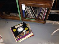 Photo showing vinyl records stored vertically on a wooden shelf, with some records scattered on the floor. Shows colorful album covers and wear.