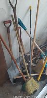 Photo showing an assortment of long handled shovels and brooms resting against a wall.