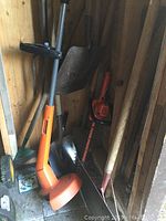 Photo showing weed eater/trimmer, shovel, rake, long-handled tool, and smaller garden tools inside a wooden shed corner.