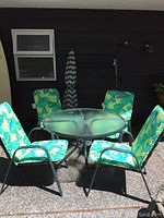 Full outdoor patio set showing glass top table, four chairs with cushions, and closed umbrella