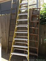 Photo showing three ladders: one large yellow metal ladder and two wooden ladders leaning against fence